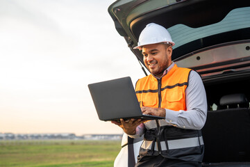 Smart engineer wearing safety uniform working with laptop sitting back of the car near airport at...