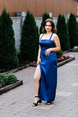 Girl in a long blue dress on the street in summer, green bushes behind her, she stands on the path