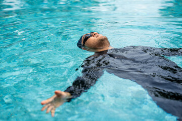 A person in a black rash guard and swim cap swims laps in a clear blue pool, demonstrating an...