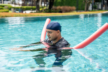 A smiling person in a swim cap and rash guard is in a pool, holding a pink swim noodle in an arc above their head. This image depicts aquatic exercise and joyful water recreation.