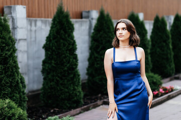 Girl in a long blue dress on the street in summer, green bushes behind her, she stands on the path