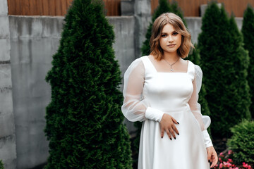 Girl in white dress on street in summer, green bushes behind her Girl looking straight into camera