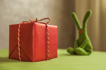 Red gift box with decorative rope bow in foreground with blurred green crochet bunny sitting in background on table