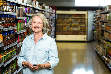 Senior woman holding smartphone and checking product info in supermarket aisle of condiment shelves
