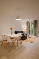 Wide shot of minimalist living and dining area with white dining table, gray sofa, floor lamp and large window