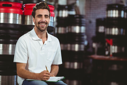 Male brewery technician sitting on stool in storage area taking clipboard notes by stainless kegs - Powered by Adobe