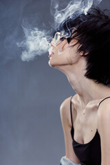 young woman wearing glasses exhaling smoke in a dark moody setting with a blue-gray background and...