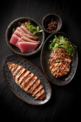 High-angle view of assorted grilled and sliced meats, including tuna and chicken, served on dark plates.  Small bowls of greens and a condiment are also visible