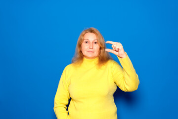 Caucasian woman in her 40s wearing a yellow sweater, smiling and confident, gesturing with her hand, making a small sign with her fingers, looking at the camera. Isolated over blue background.