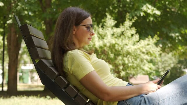 A woman in a yellow shirt sits on a wooden bench reading from a tablet. She enjoys a peaceful moment surrounded by lush greenery in a sunlit park in Simferopol, Crimea.