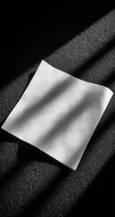 A square sheet of white paper lies on a dark surface, illuminated by angled sunlight casting dramatic shadows