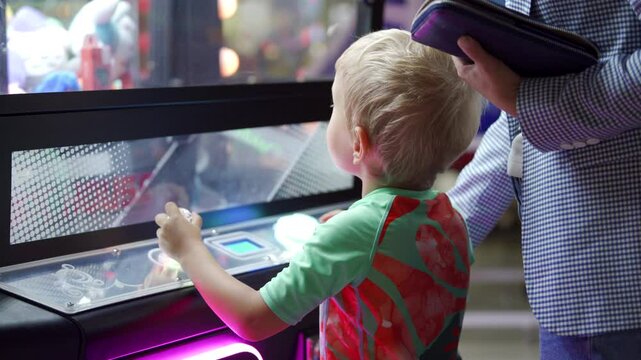 A young boy intently operates the joystick of a claw machine while standing next to a supportive adult. They are inside a vibrant indoor amusement arcade.