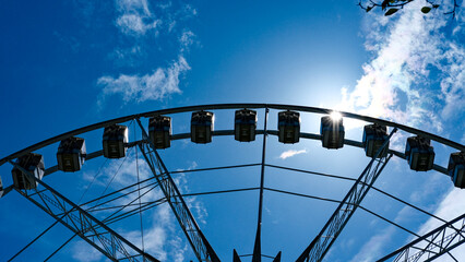 Ferris wheel photographed from below against a blue sky with sun flare and scattered clouds.