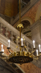 Golden chandelier with candle-like bulbs hanging in an opulent classical interior with stone railings.