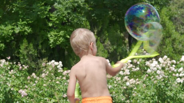 A young boy in orange shorts swings a yellow bubble wand, forming large iridescent bubbles. The vibrant greenery and blooming flowers in Simferopol, Crimea, provide a joyful and lively outdoor setting