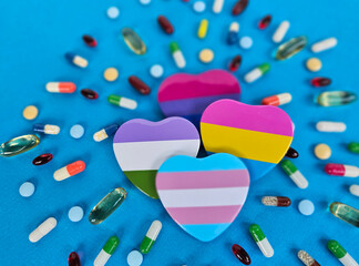 Colorful heart shapes symbolizing inclusivity are arranged amidst different types of pills on a bright blue surface, showcasing a blend of health