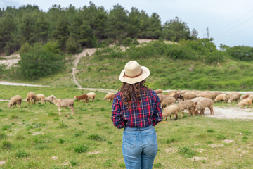 A woman wearing a straw hat stands in a field with a herd of sheep
