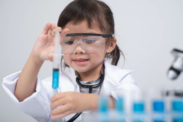 A young girl in a lab coat and safety goggles is carefully mixing liquids in a laboratory setting,...