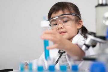 A young child in a lab coat and safety goggles is carefully holding a test tube filled with blue...