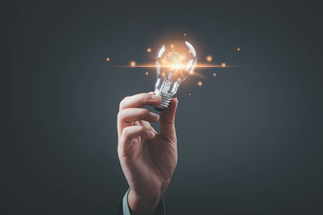 A hand holding a glowing light bulb, symbolizing ideas, inspiration, and innovation against a dark...