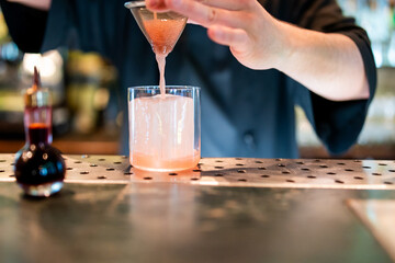 Bartender straining pink cocktail into a glass on a perforated metal bar counter, with bottle in...