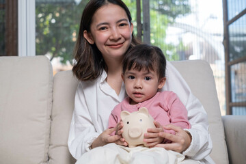 A mother and her child sit together on a couch, holding a piggy bank, symbolizing savings and...