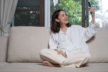 A young woman sits comfortably on a sofa, smiling while taking a selfie, surrounded by natural...