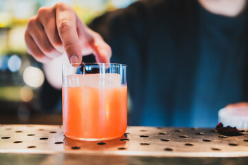 Close-up of a bartender’s hand placing garnish into an orange cocktail on a perforated metal bar...