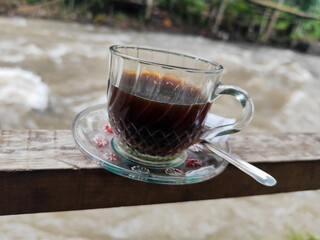 A cup of hot black coffee in a glass cup with matching saucer and spoon, placed on the edge of a...