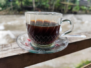 A cup of hot black coffee in a glass cup with matching saucer and spoon, placed on the edge of a...