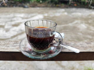 A cup of hot black coffee in a glass cup with matching saucer and spoon, placed on the edge of a...