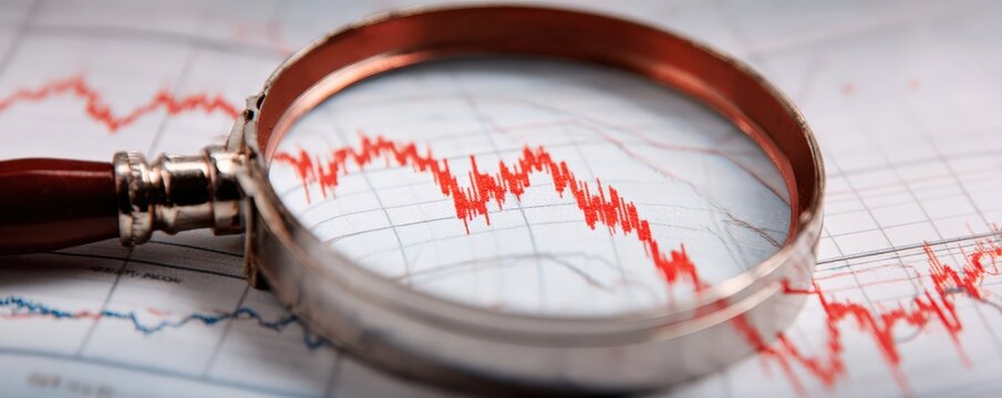 A magnifying glass closely examines a downward-trending red financial chart line on a graph paper background.