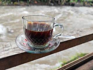 A cup of hot black coffee in a glass cup with matching saucer and spoon, placed on the edge of a...