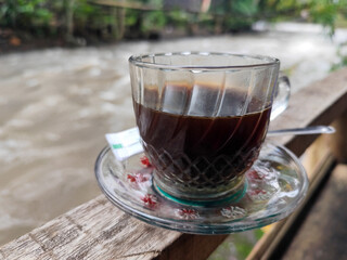 A cup of hot black coffee in a glass cup with matching saucer and spoon, placed on the edge of a weathered wooden fence with a backdrop of a flowing river and blurry green trees. Outdoor cafe menu.