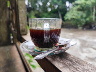 A cup of hot black coffee in a glass cup with matching saucer and spoon, placed on the edge of a...
