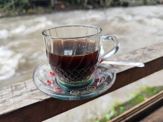 A cup of hot black coffee in a glass cup with matching saucer and spoon, placed on the edge of a weathered wooden fence with a backdrop of a flowing river and blurry green trees. Outdoor cafe menu.