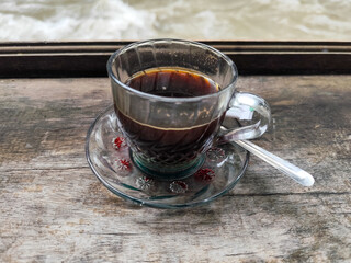 A cup of hot black coffee in a glass cup with matching saucer and spoon, placed on the edge of a weathered wooden fence with a backdrop of a flowing river and blurry green trees. Outdoor cafe menu.