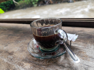 A cup of hot black coffee in a glass cup with matching saucer and spoon, placed on the edge of a weathered wooden fence with a backdrop of a flowing river and blurry green trees. Outdoor cafe menu.