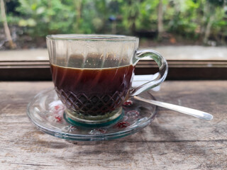 A cup of hot black coffee in a glass cup with matching saucer and spoon, placed on the edge of a...