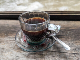 A cup of hot black coffee in a glass cup with matching saucer and spoon, placed on the edge of a weathered wooden fence with a backdrop of a flowing river and blurry green trees. Outdoor cafe menu.
