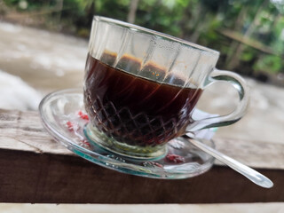 A cup of hot black coffee in a glass cup with matching saucer and spoon, placed on the edge of a...