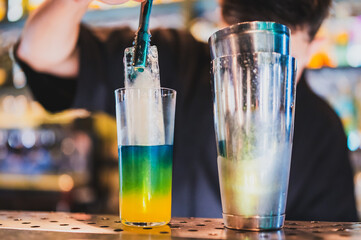 Bartender prepares a colorful layered cocktail with ice using tongs, beside a metal shaker on a bar...