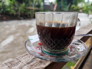 A cup of hot black coffee in a glass cup with matching saucer and spoon, placed on the edge of a...