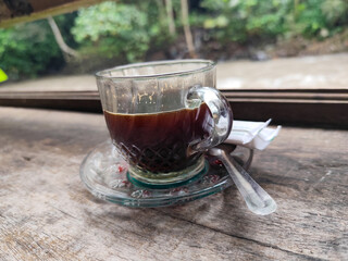 A cup of hot black coffee in a glass cup with matching saucer and spoon, placed on the edge of a...