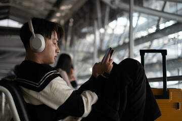 A young man with headphones sits in an airport, looking at his phone, embodying tranquility amidst...