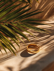 Obraz premium Golden Bracelet in Sunlight: An elegant gold bracelet rests gracefully amidst the shadows of a tropical palm leaf, casting intricate patterns on a warm, textured surface.