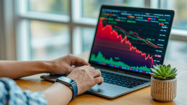 Close-up of hands using MacBook showing red stock graph, with plant nearby on wooden desk, natural light, blurred background, warm tones throughout