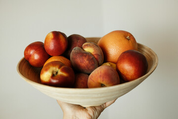 Hand holding a wooden bowl full of fresh summer fruits - nectarines, peaches, oranges, and grapefruit against a neutral indoor background.