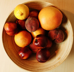 Top-down view of a wooden bowl filled with citrus and stone fruits: lemons, oranges, nectarines, and peaches. Natural warm tones and soft lighting