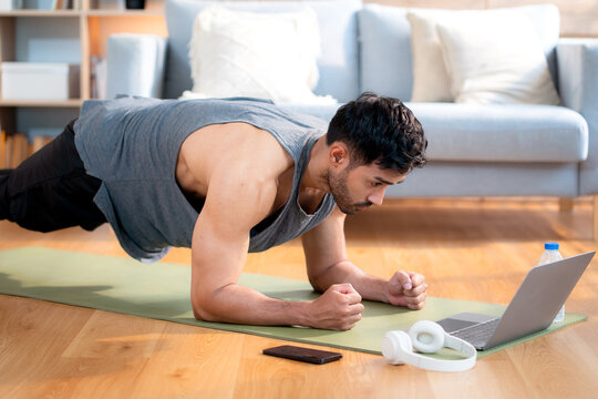Young asian man doing plank exercise in living room at home while watching online workout tutorial on laptop with endurance and motivation, man training plank on yoga mat, digital fitness lifestyle.